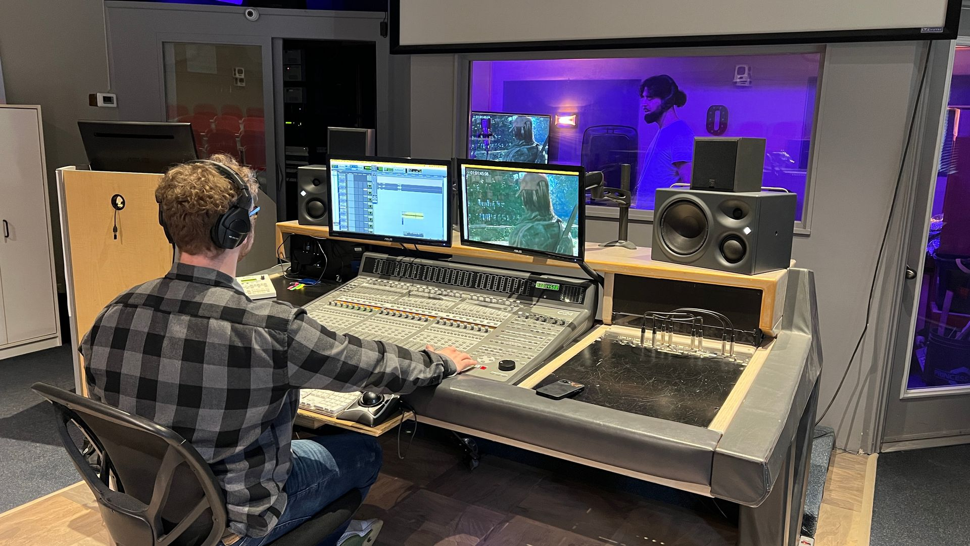 A person wearing headphones works at a sound mixing console in a studio with multiple monitors.