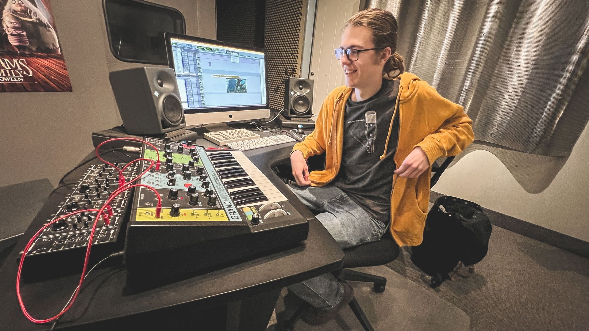 Person in yellow hoodie at a desk with computer and music equipment, smiling in a recording studio.