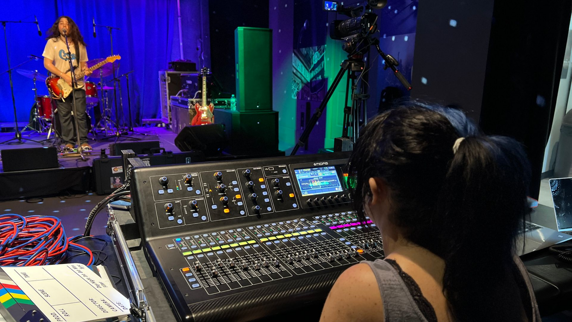 Woman at soundboard, band on stage. Stage has lighting and instruments. Green and blue hues.