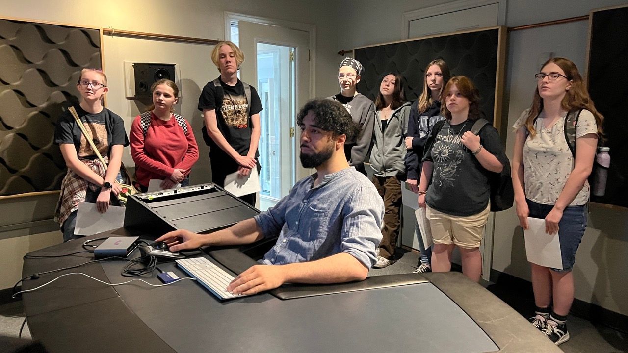 A group of people are standing around a man sitting at a desk.