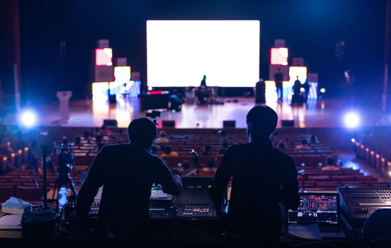 Two sound engineers are standing in front of a large screen on a stage setting up for a concert