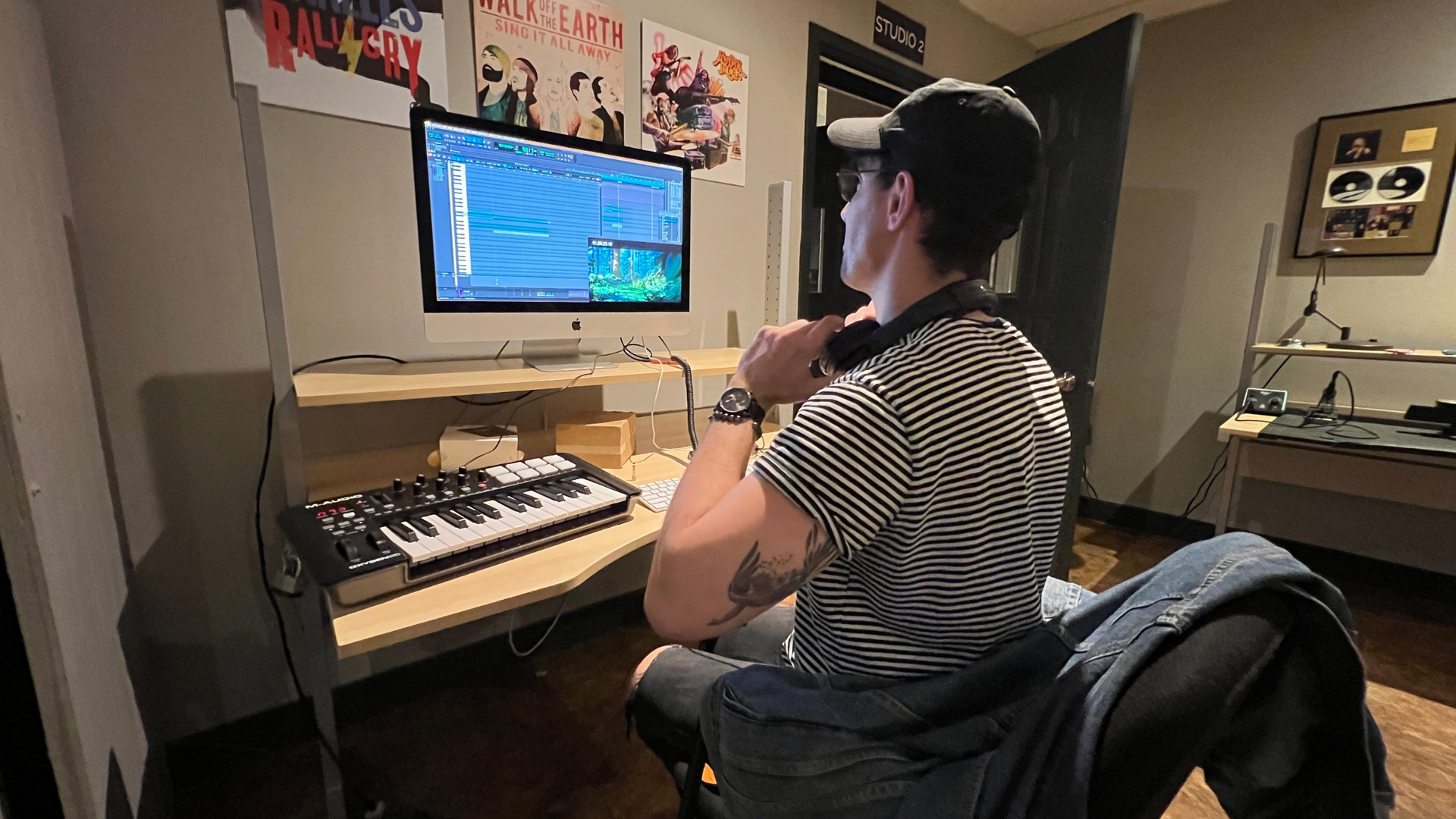 Person wearing hat at computer with keyboard, headphones, in a studio with posters.