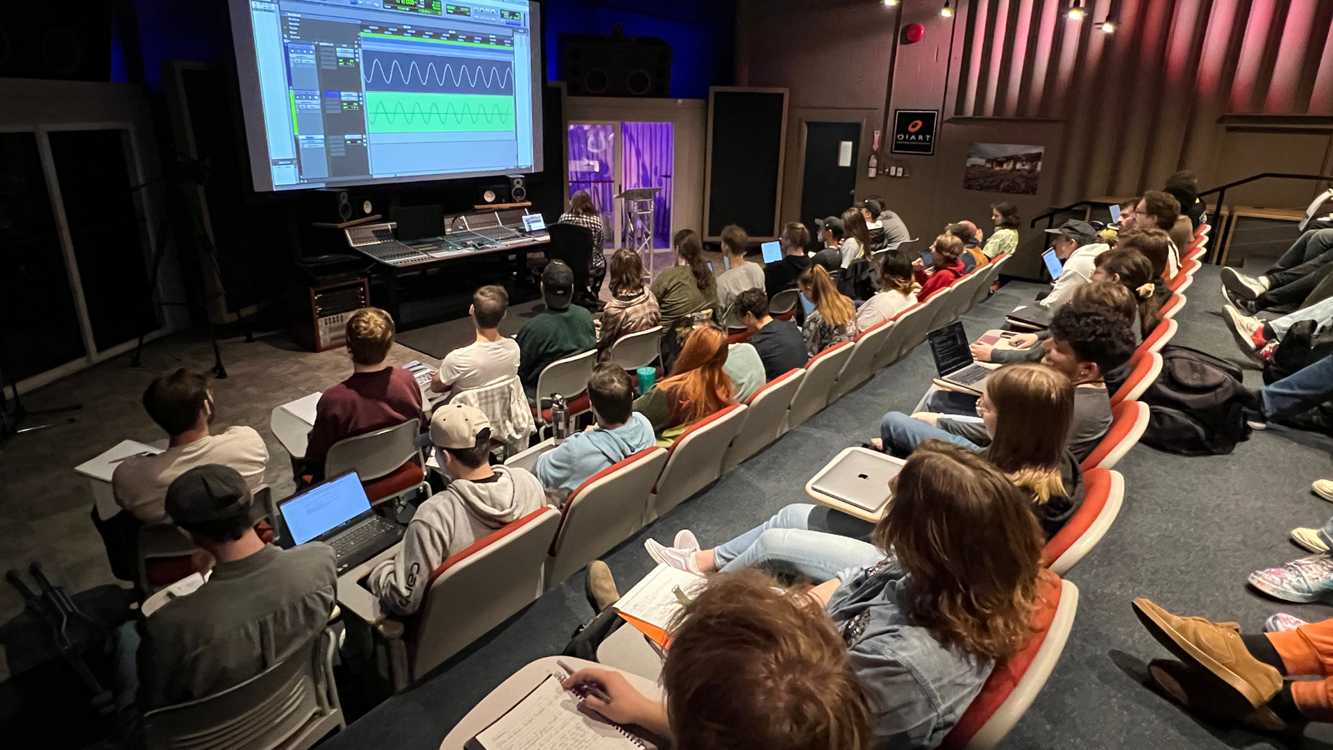 A classroom with students seated in rows facing a large projection screen displaying digital audio workstation software.