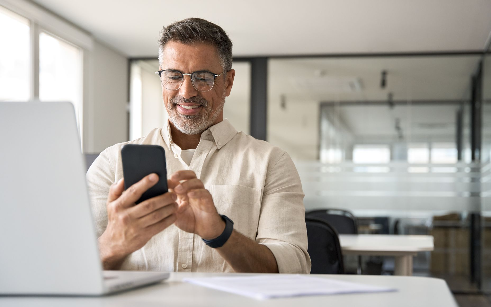 Man wearing glasses smiles while looking at a smartphone in an office setting.