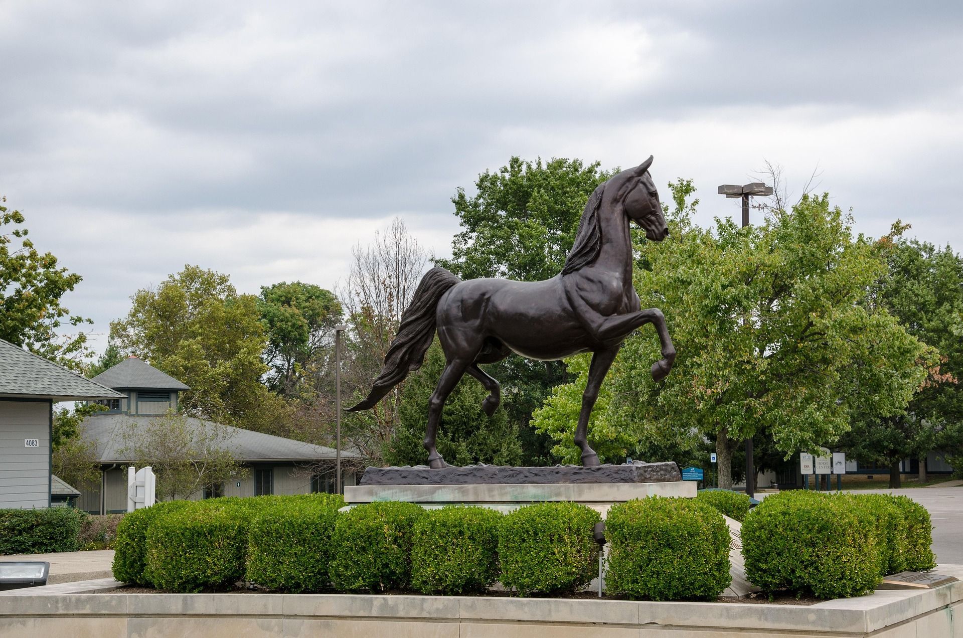 Bronze horse statue rearing on a pedestal, set against a cloudy sky and green trees.