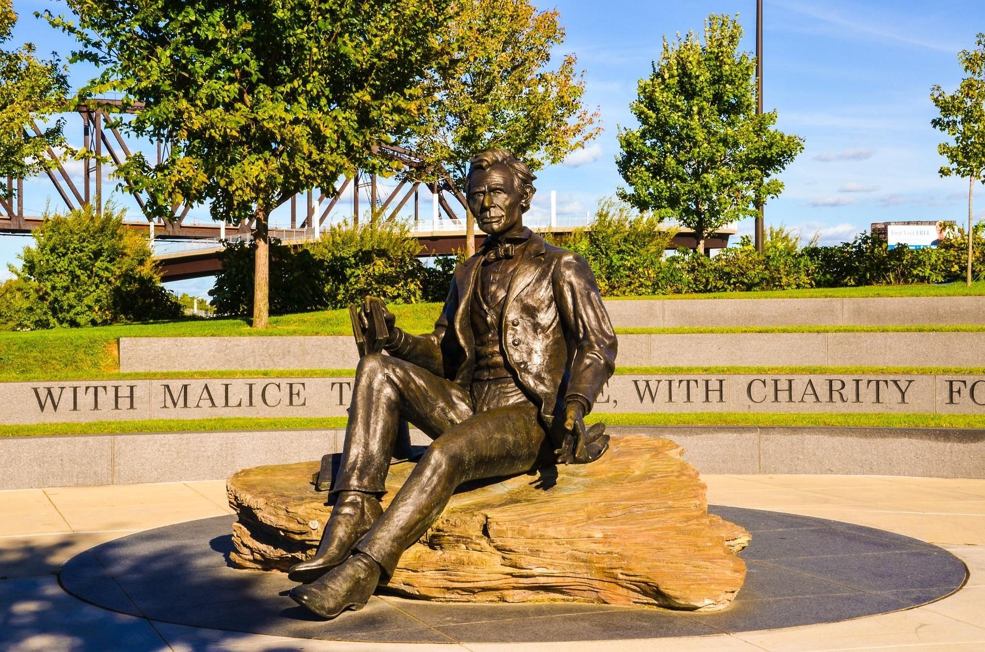 Bronze statue of a seated man holding a small object, atop a rock, set in a plaza with trees and text.