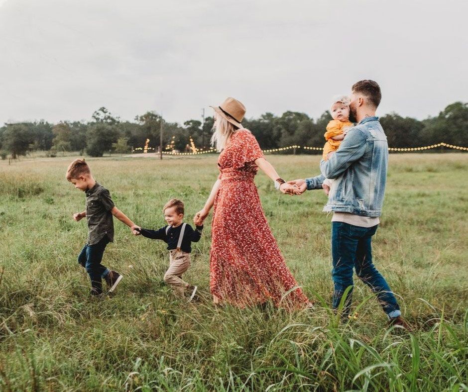 Family walks hand-in-hand through a grassy field. Two children run ahead, parents trail behind holding a baby.