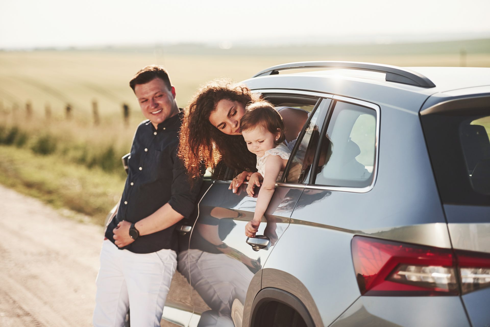Family smiles from car window in field.