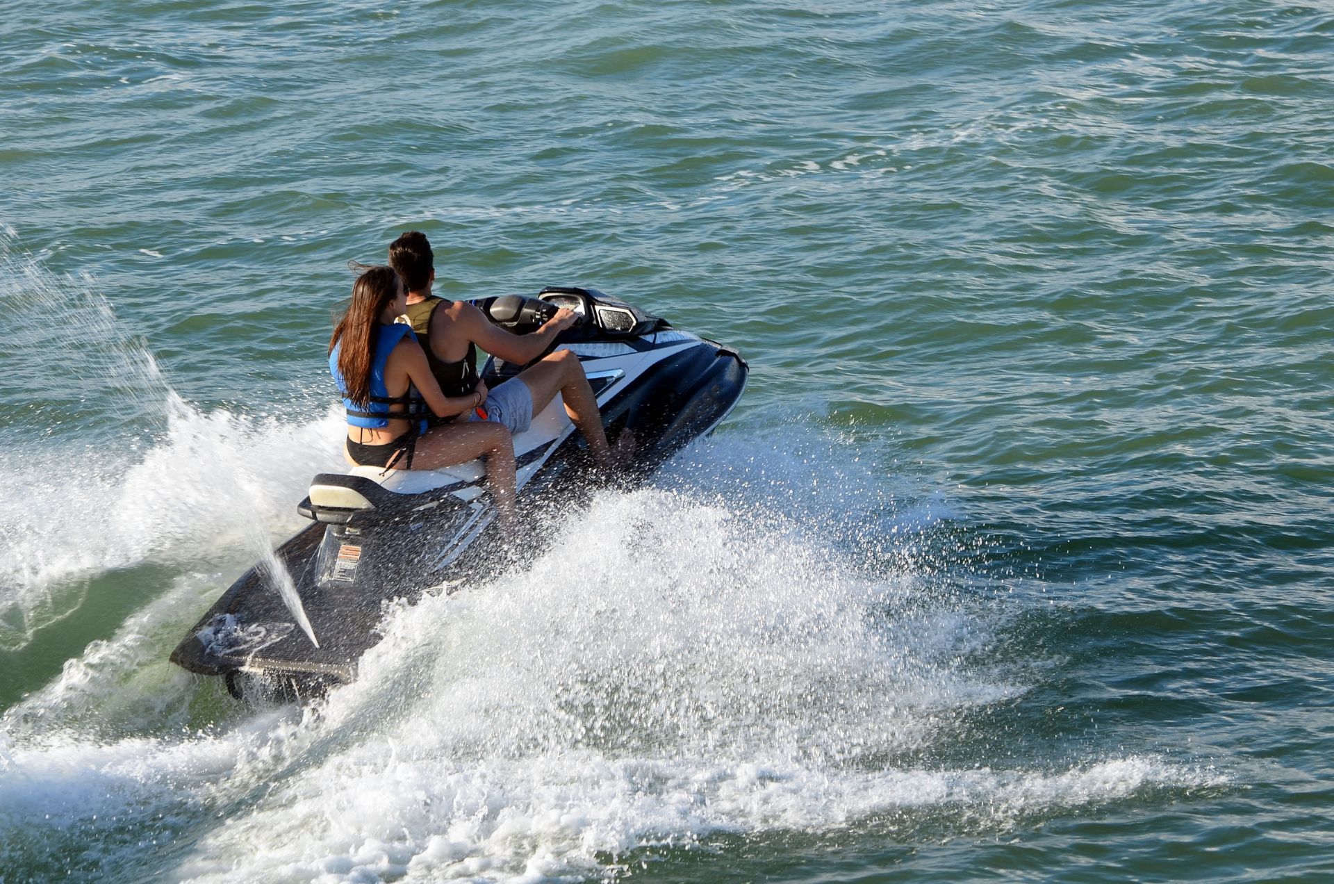 Couple on a jet ski, speeding across choppy blue water, creating white spray.