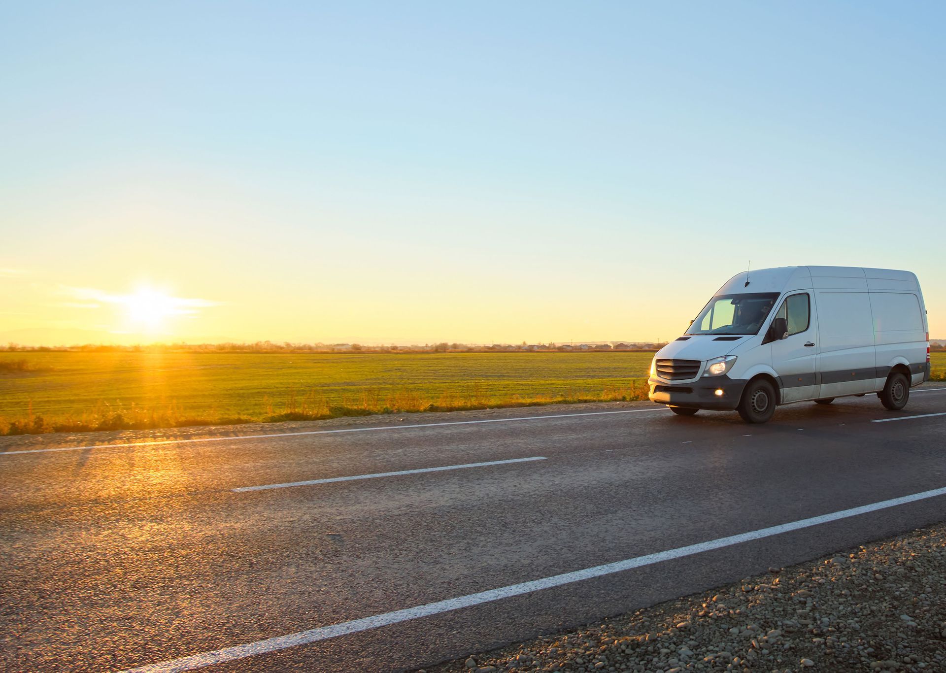 White delivery van driving on a road at sunset, with a field in the background.