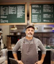 Man in apron and cap smiles in front of restaurant menu boards.