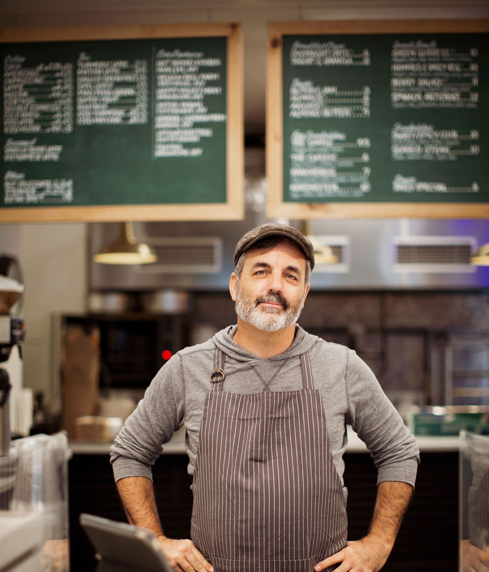 Man in apron and cap smiles in front of restaurant menu boards.