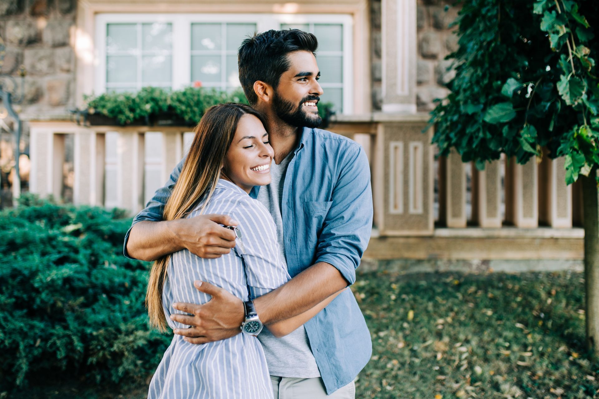 Couple embracing outside a house, smiling. Man in blue shirt, woman in striped dress, green yard.