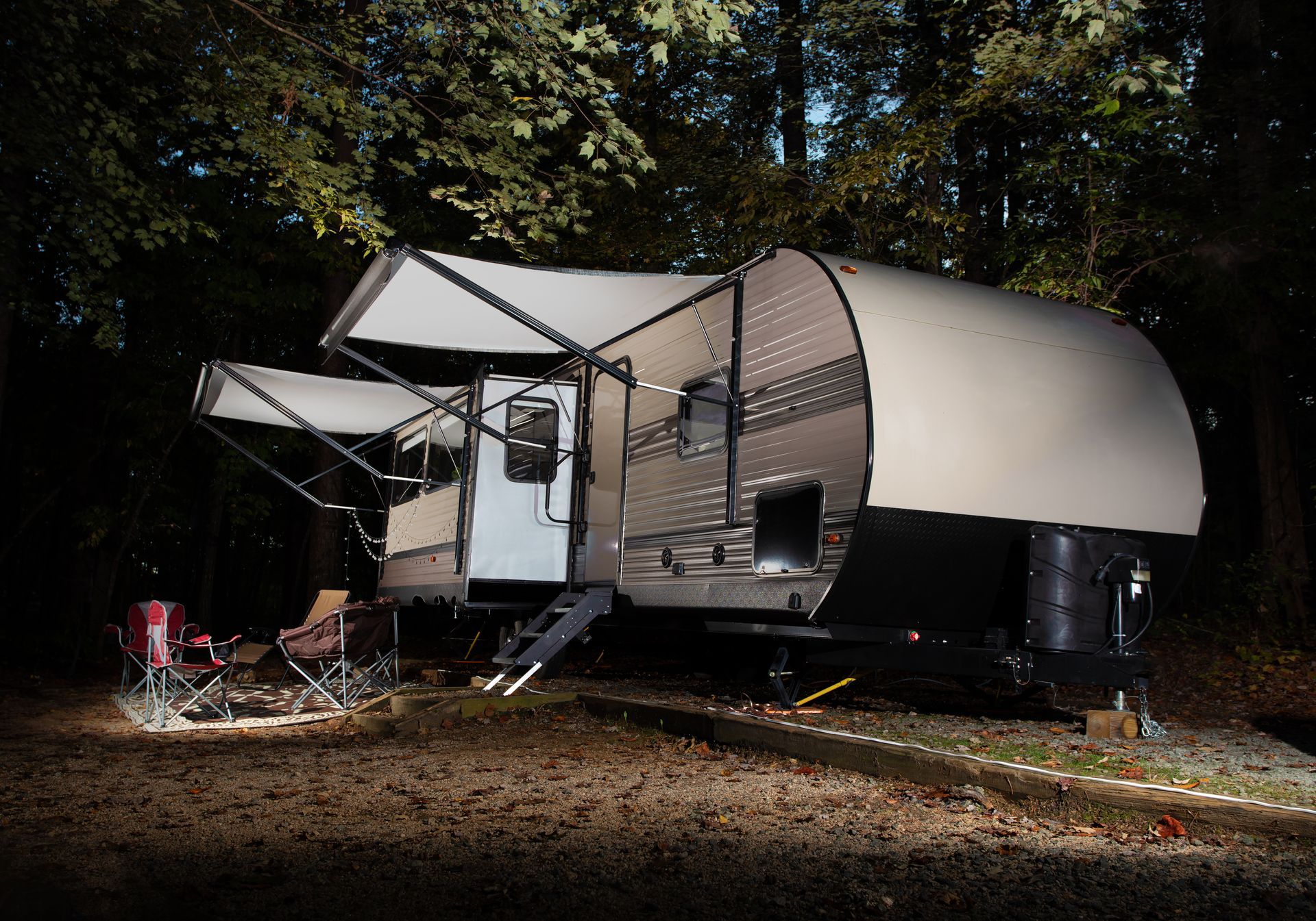 A futuristic-looking camper trailer in a wooded area at night. Awning extended, lights on.