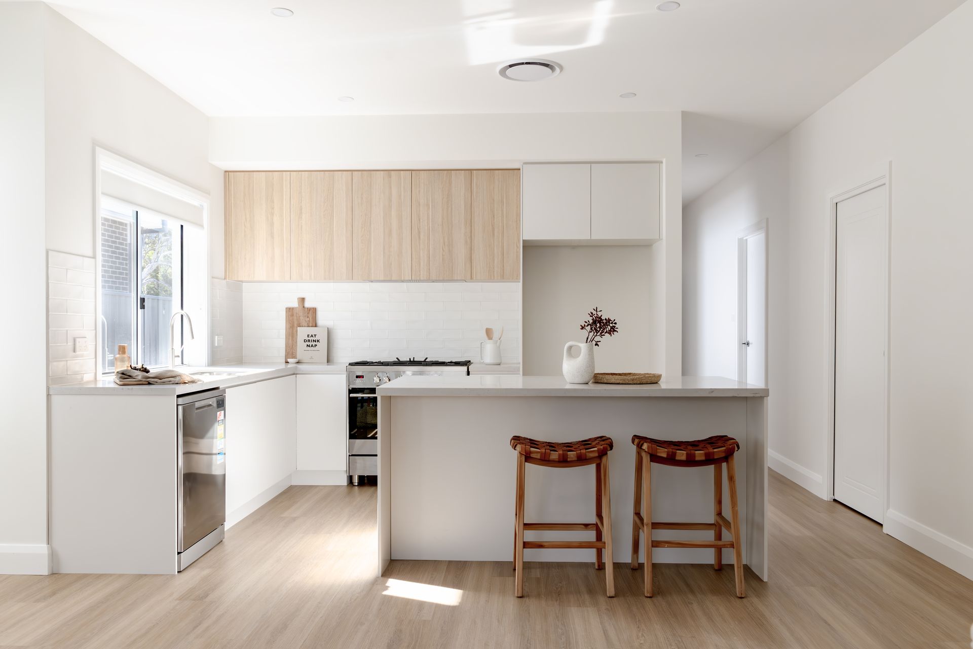 Modern White Kitchen With Light Wood Cabinets, Breakfast Bar With Two Stools, and Natural Light — Evolution Building Group in Dapto, NSW
