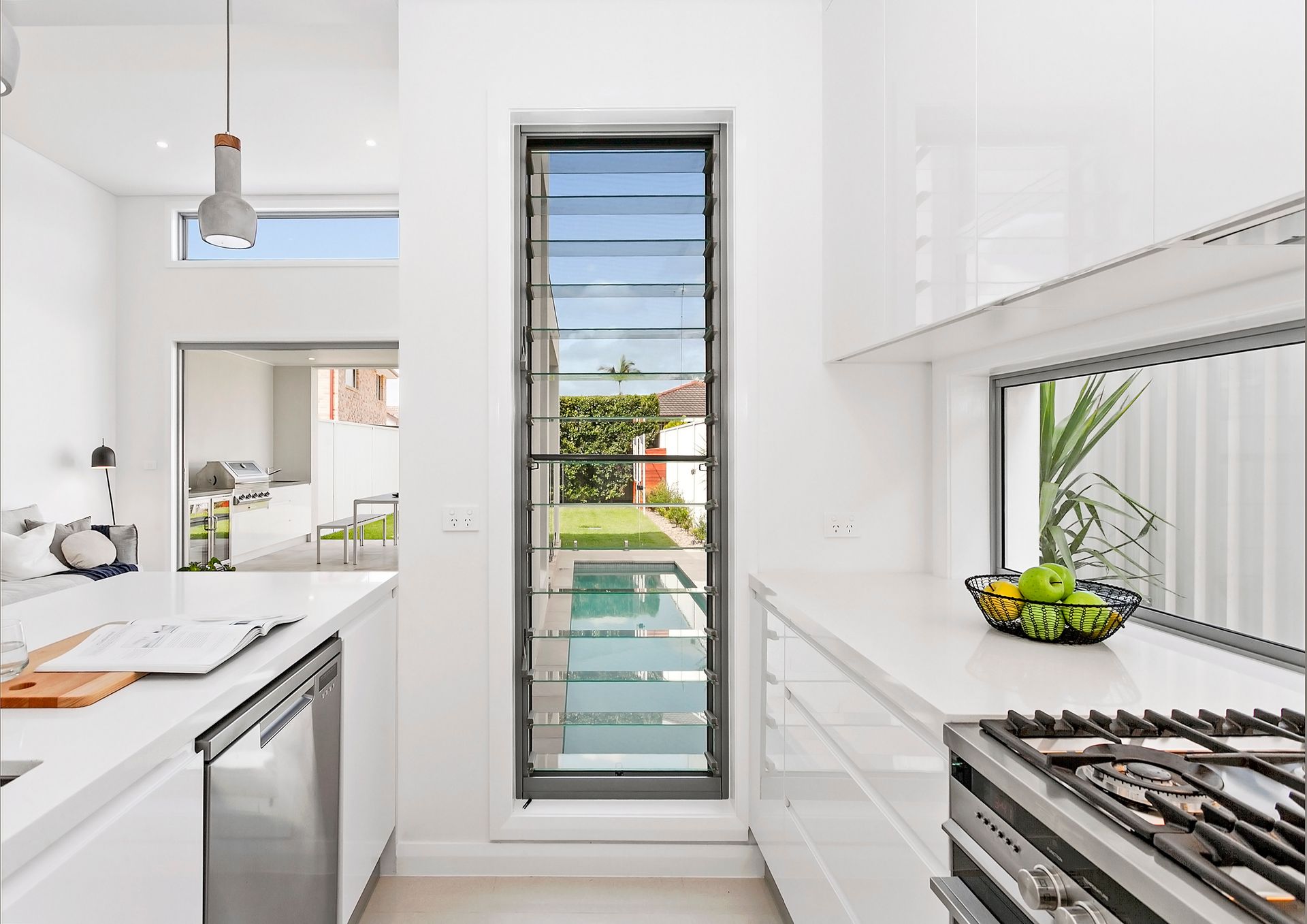 Modern kitchen with white cabinets, countertops, and a tall, narrow window overlooking a pool. A fruit bowl sits on the counter near a gas stovetop — Evolution Building Group In Moss Vale, NSW