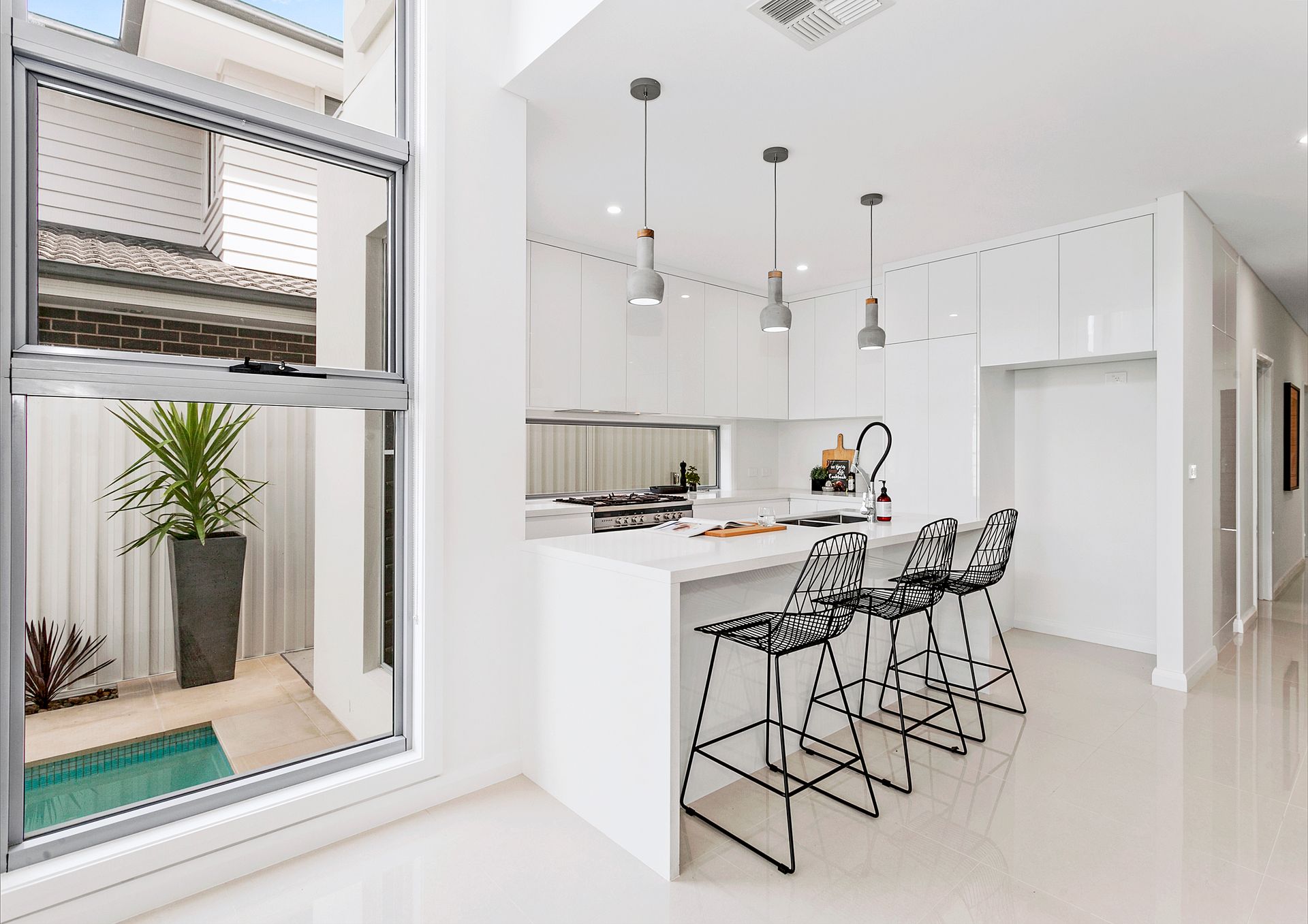 Modern, all-white kitchen with a central island and bar seating. A large window overlooks a small courtyard with a pool — Evolution Building Group In Calderwood, NSW