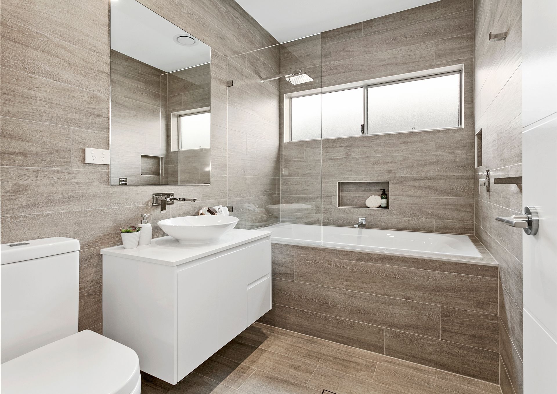 Modern bathroom with neutral stone tile walls, white vanity and toilet, and a built-in bathtub — Evolution Building Group In Appin, NSW