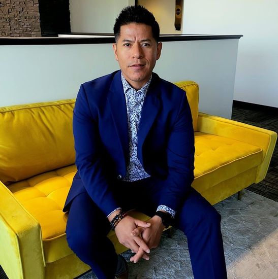 Person working at a wooden desk with laptop, plant and vase. Light-colored chair, rug, and stool in a white room.