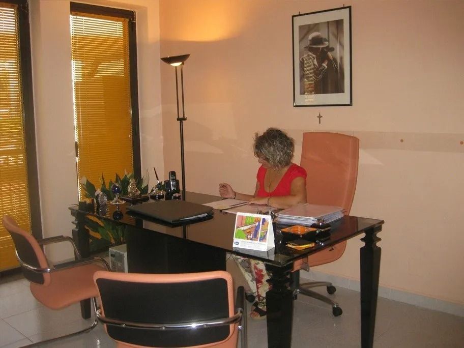 Woman at desk in office, writing. Desk has laptop and files. Tan walls, orange accents.
