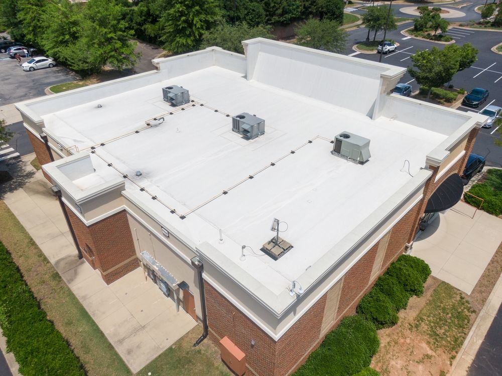 Aerial view of a white-roofed brick building with rooftop HVAC units and surrounding trees and parking lot