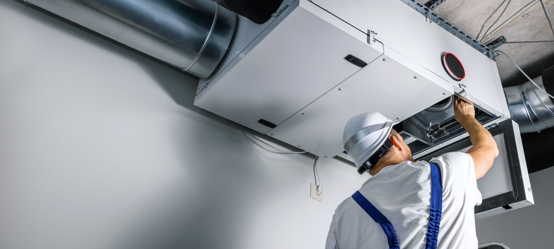 An HVAC technician in a white hard hat and work overalls inspects an air handler on a ceiling.