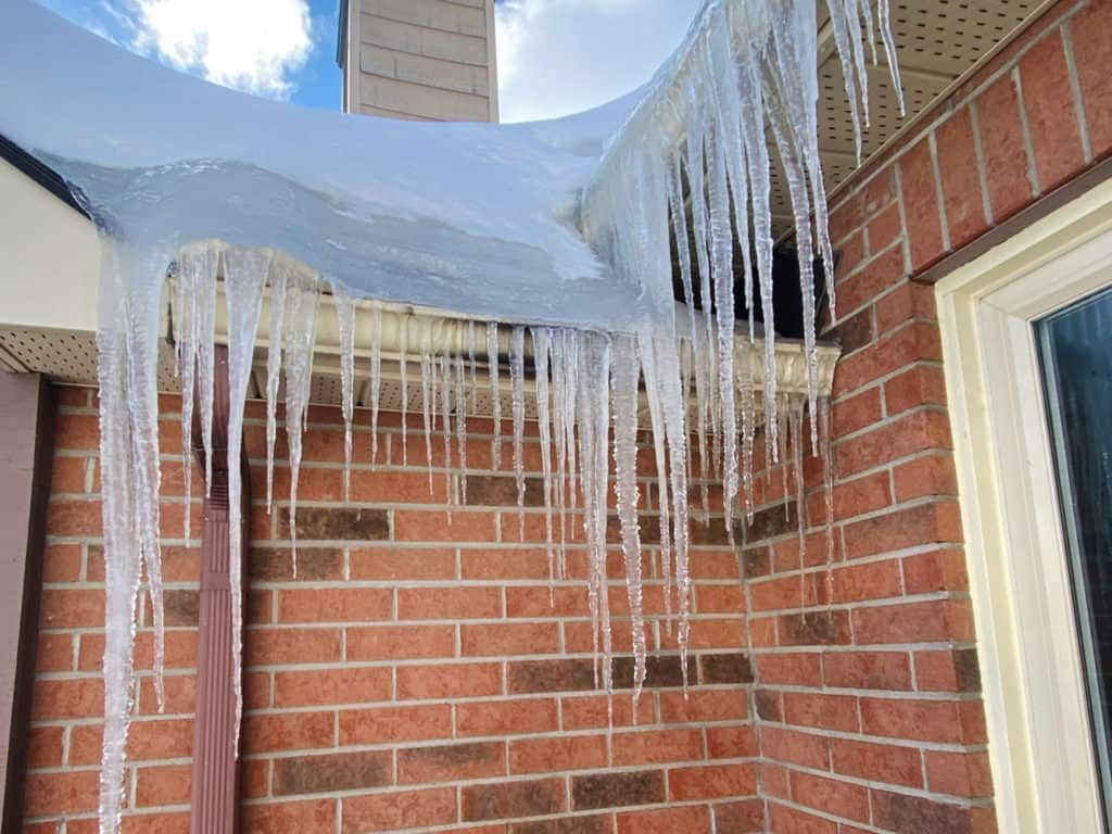 Large icicles hanging from a roof's edge, over brick and a window, with snow on top.