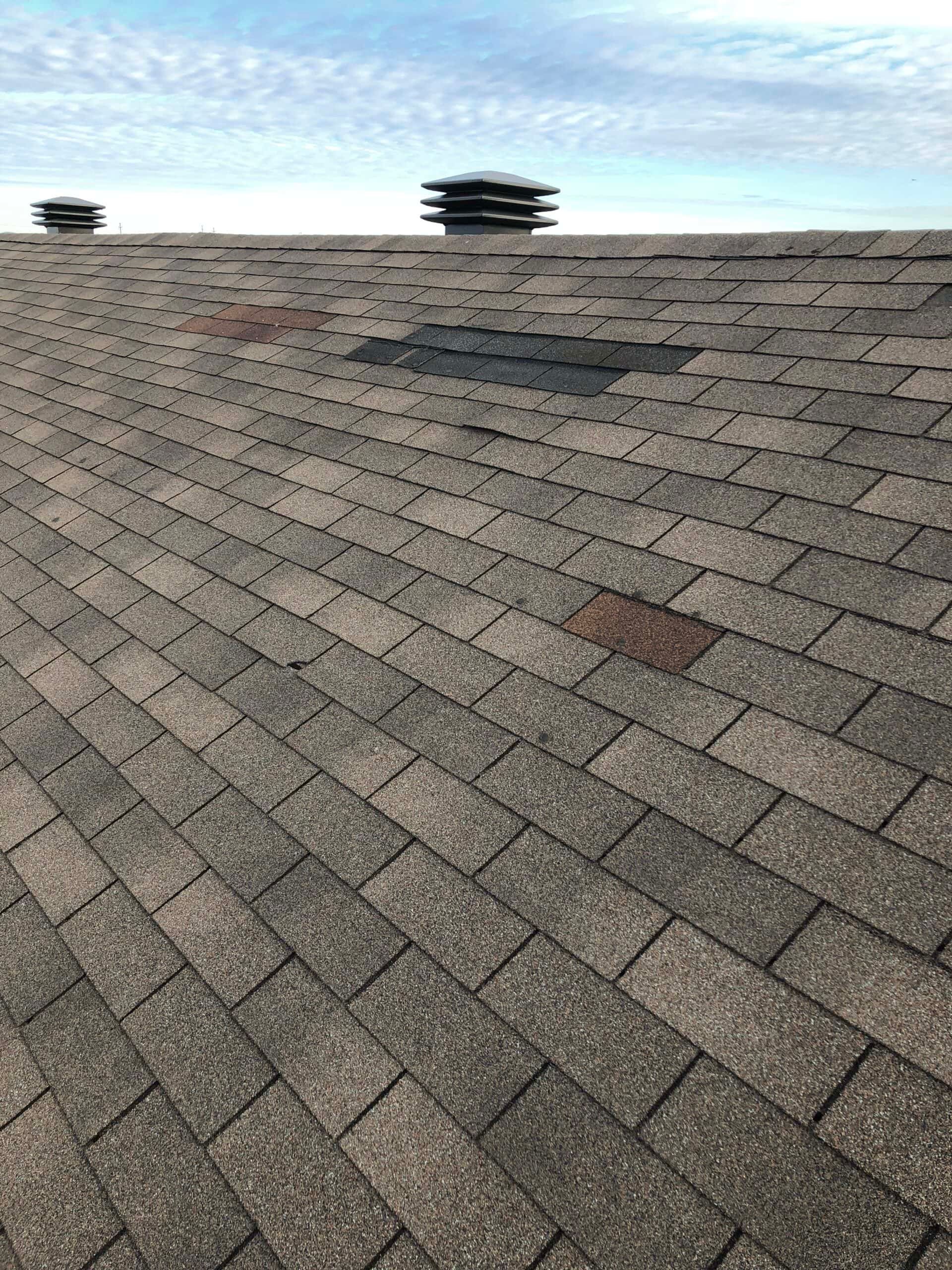 Damaged asphalt shingle roof with missing and discolored shingles, two vents, and a cloudy sky.