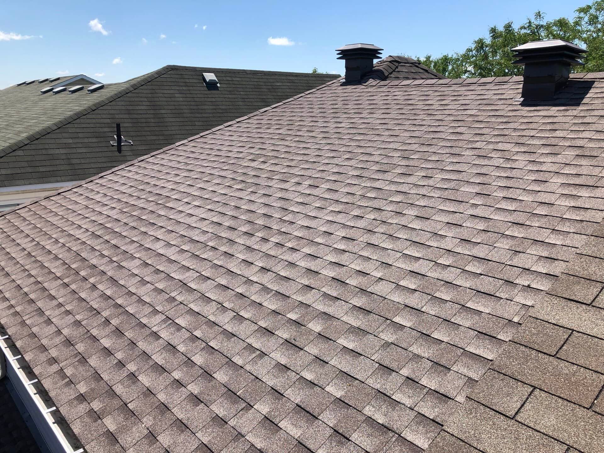 Brown shingle roof with two chimneys against a blue sky.