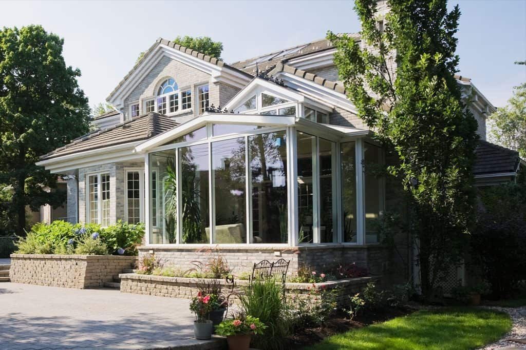 Sunroom addition on a two-story house with glass walls and a stone facade. Lush greenery surrounds the home.