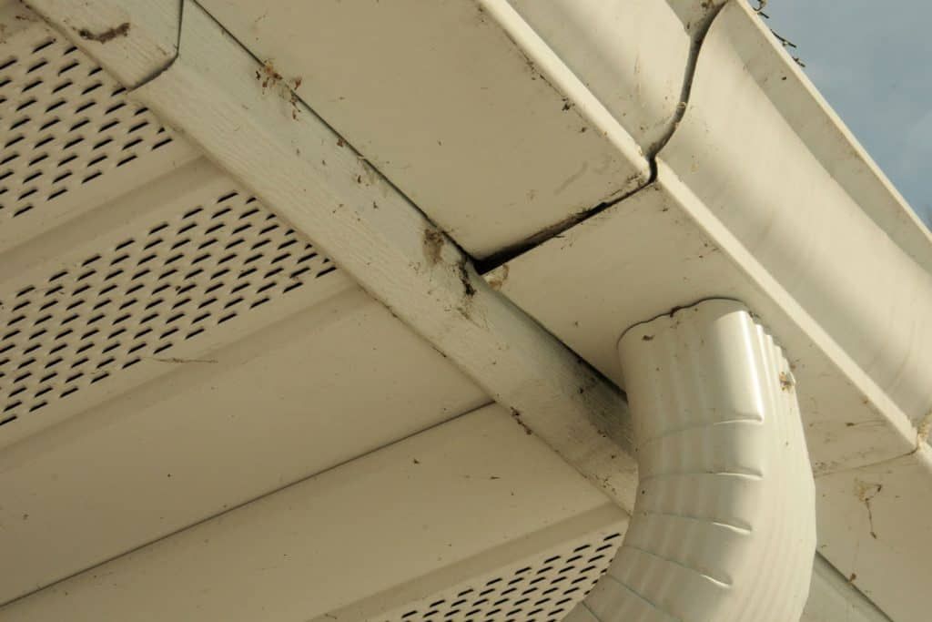 White gutter system with a downspout, showing a cracked seam and debris.