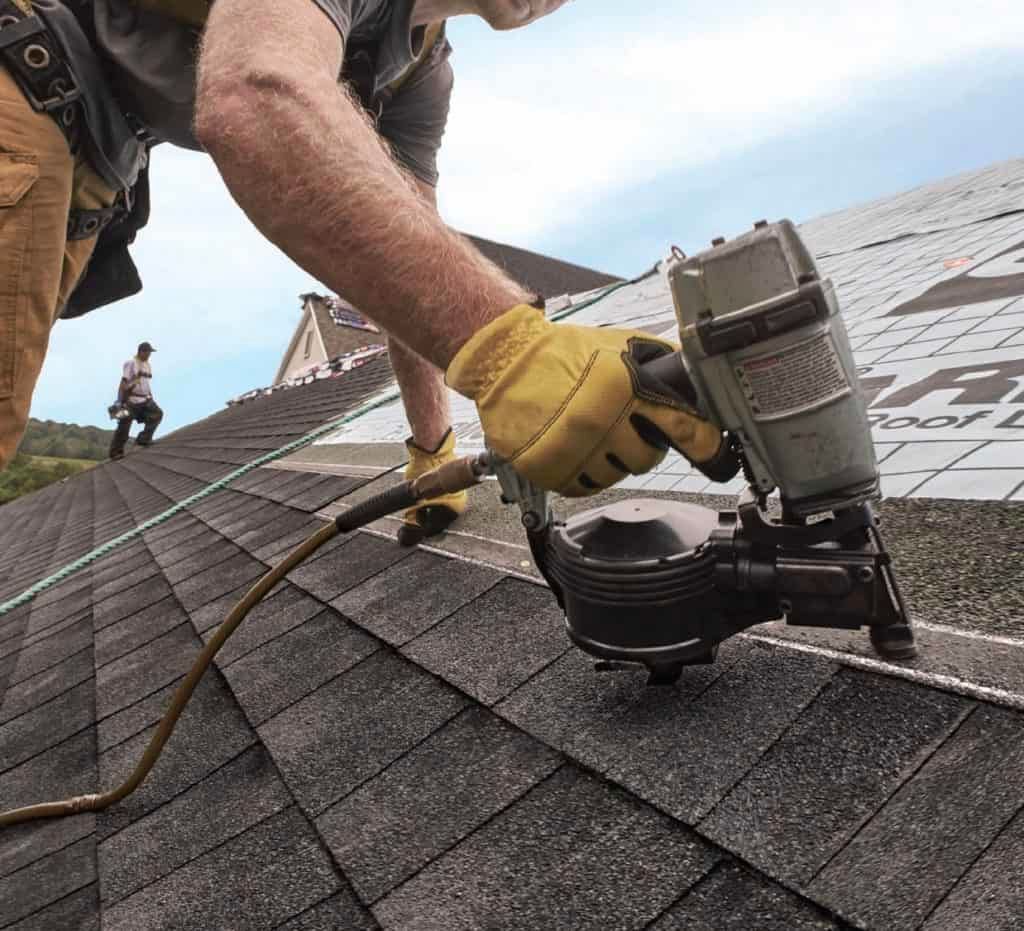 Roofer using a nail gun to install shingles on a roof. Yellow gloves and a second worker in the background.