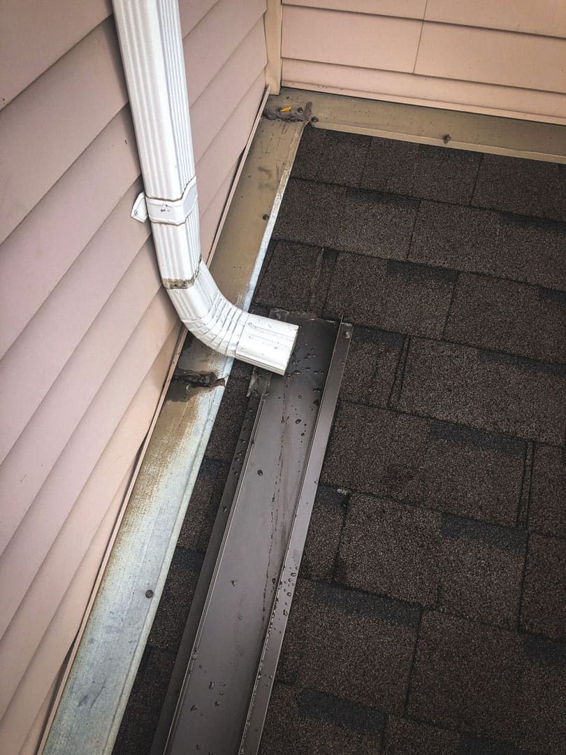 Gutter connected to a downspout on a roof corner. The gutter is filled with debris and has water stains.