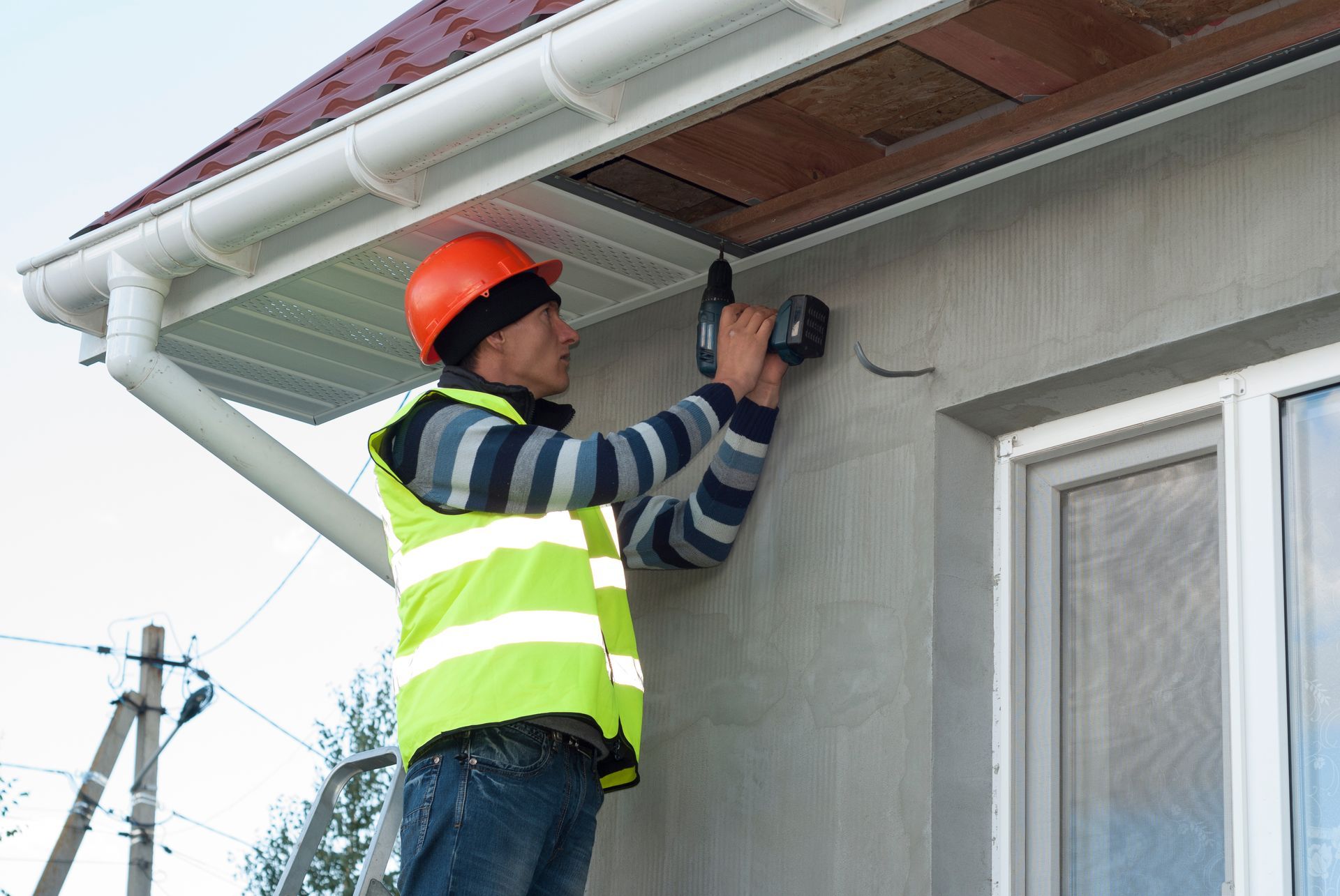 Person in safety vest and hard hat using a drill on a building's exterior.