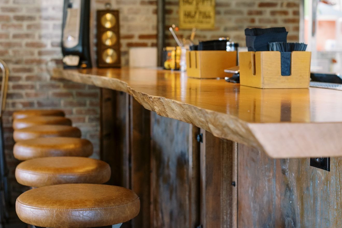 A long wooden bar with stools in a restaurant with a brick wall.