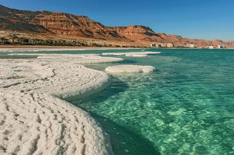 Turquoise water laps against white salt deposits at the shore of the Dead Sea, with arid mountains in the background.
