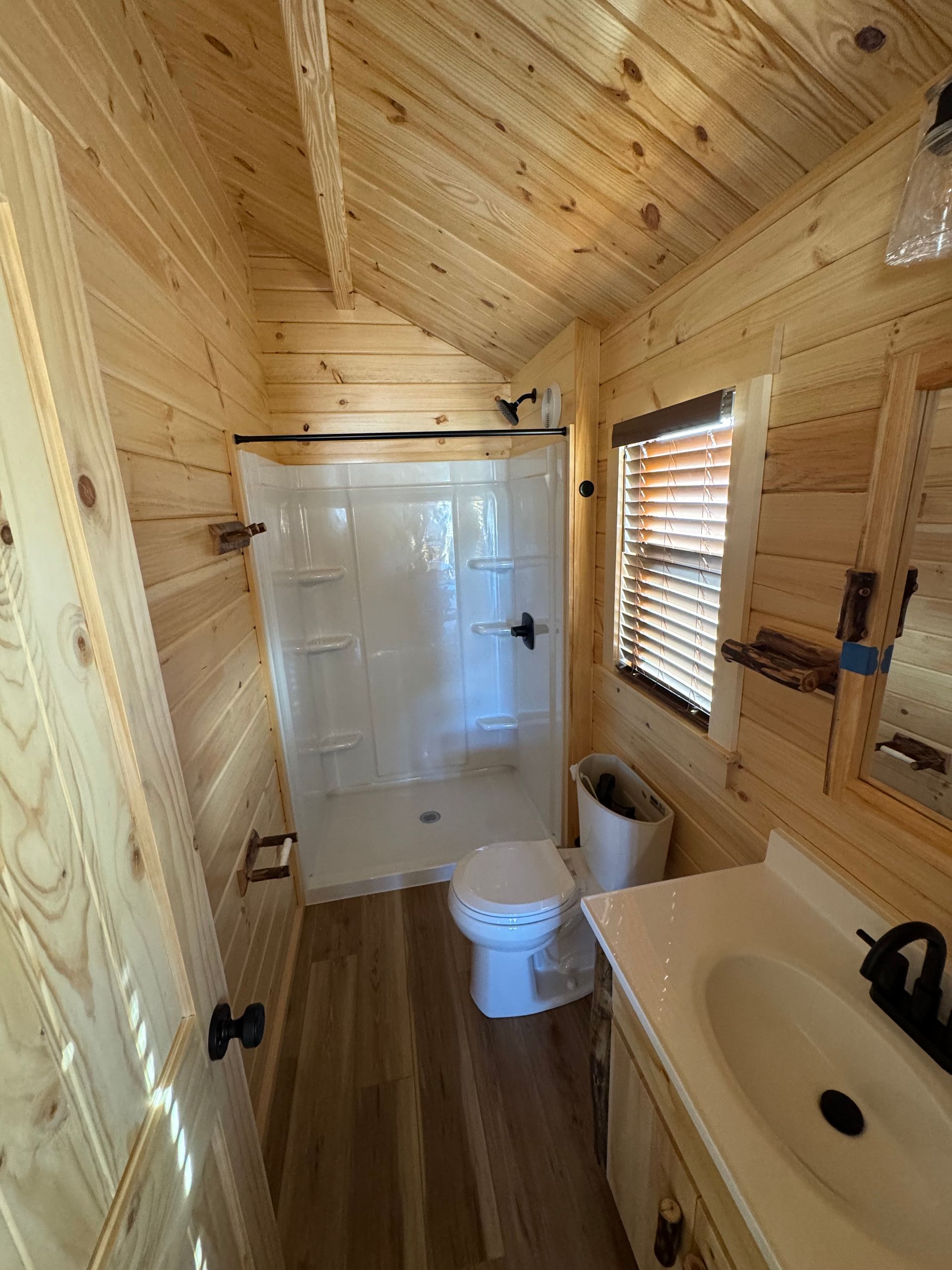 A small, rustic bathroom with light-wood paneled walls, a corner shower, a toilet, and a vanity with a white sink.