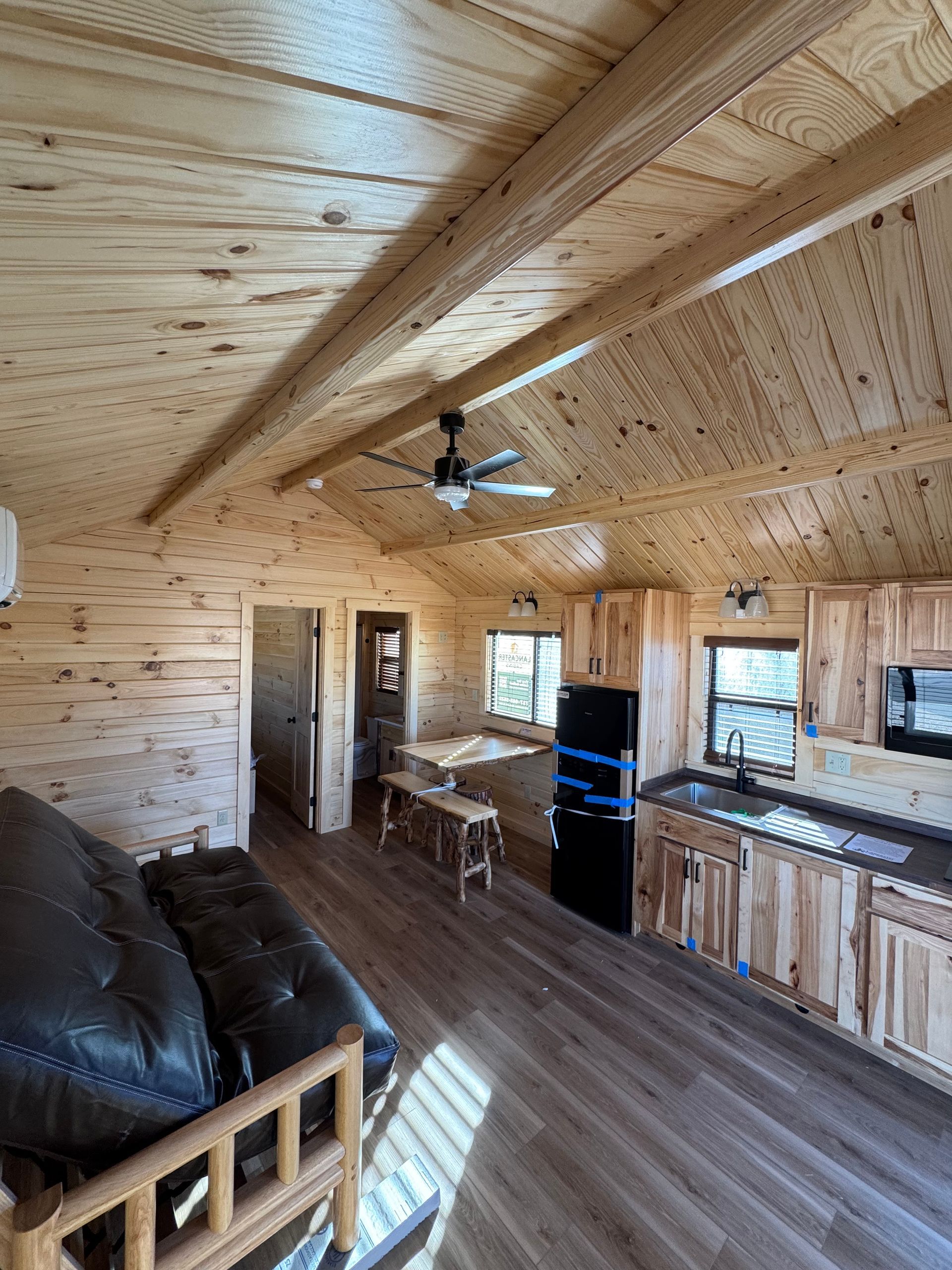 A rustic, wooden-walled cabin interior featuring a kitchenette, a black futon, a small table, and a ceiling fan.
