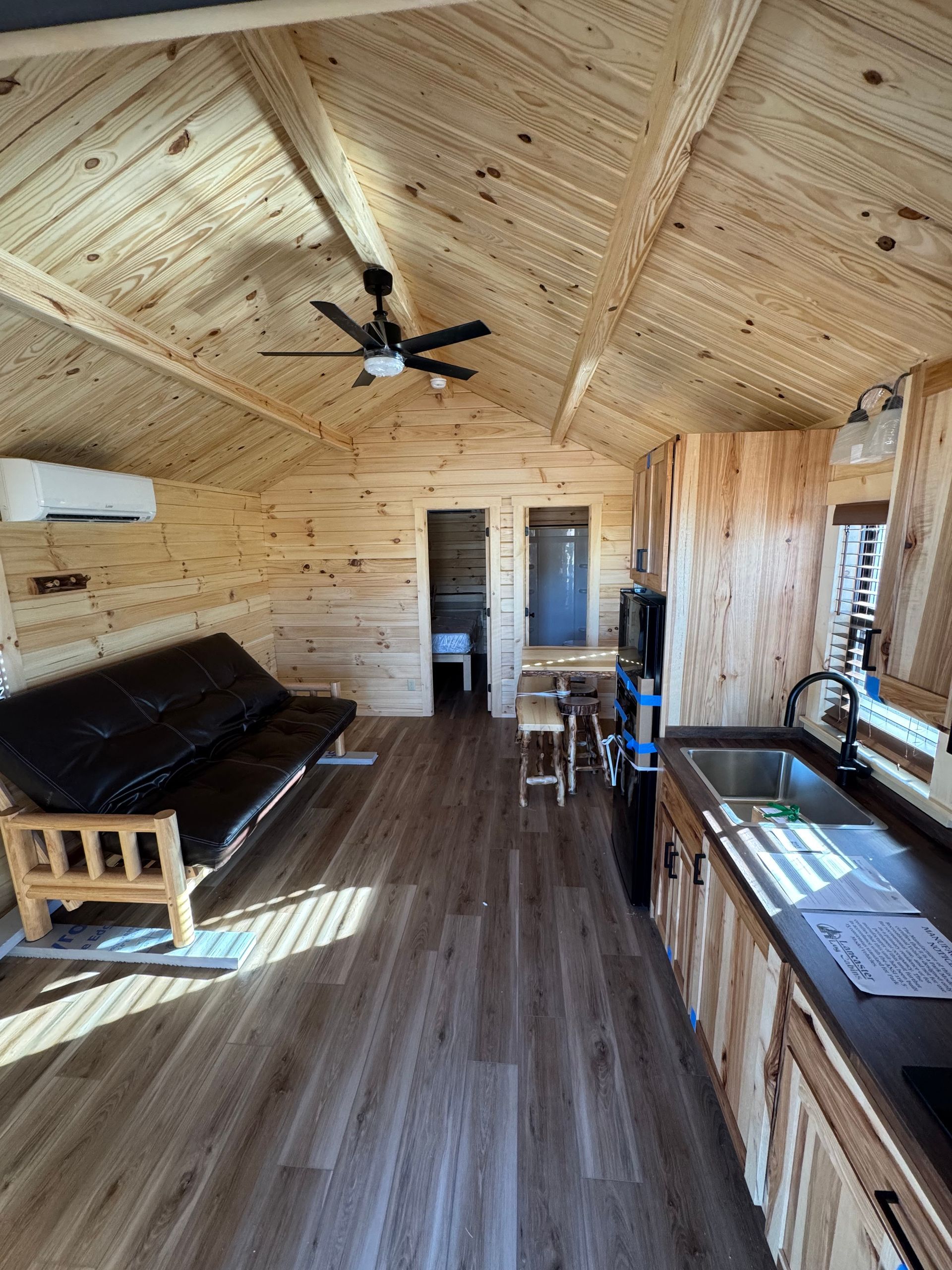 Interior of a small rustic cabin with wood-paneled walls, a black futon, kitchenette, and a ceiling fan.