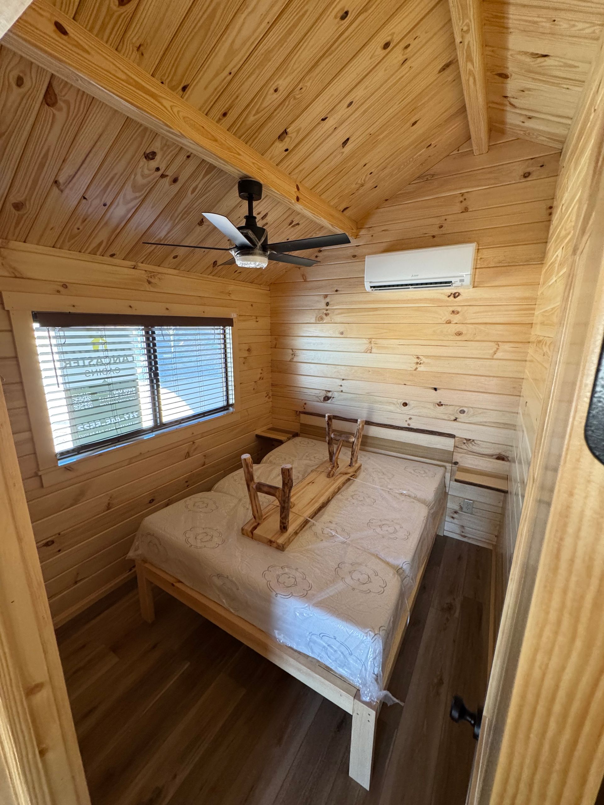 A small cabin bedroom with light pine walls, a double bed with a wooden tray, a ceiling fan, and a wall-mounted AC unit.