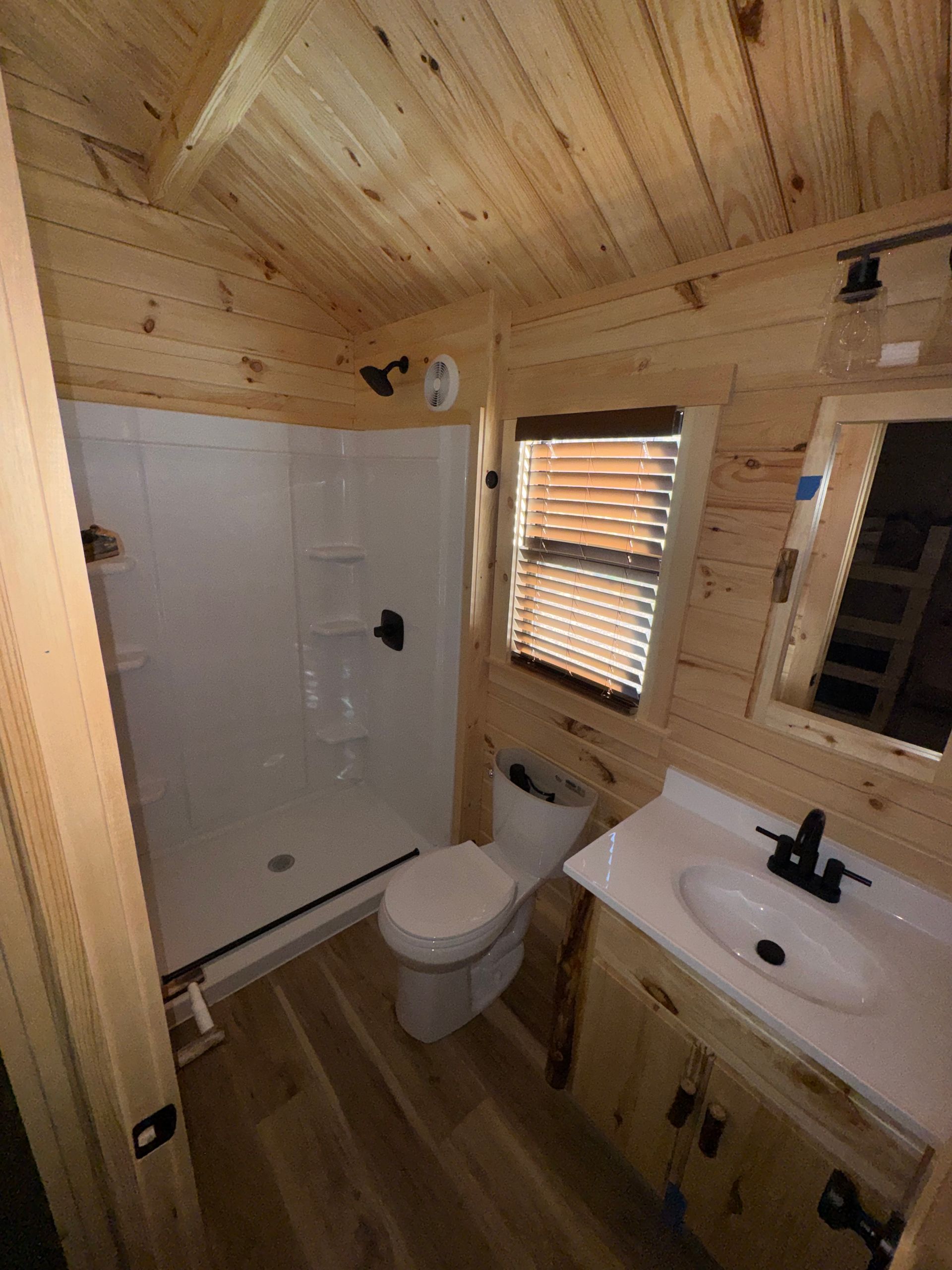 A small, rustic bathroom featuring light wood paneling, a white shower stall, a toilet, and a vanity with a sink.