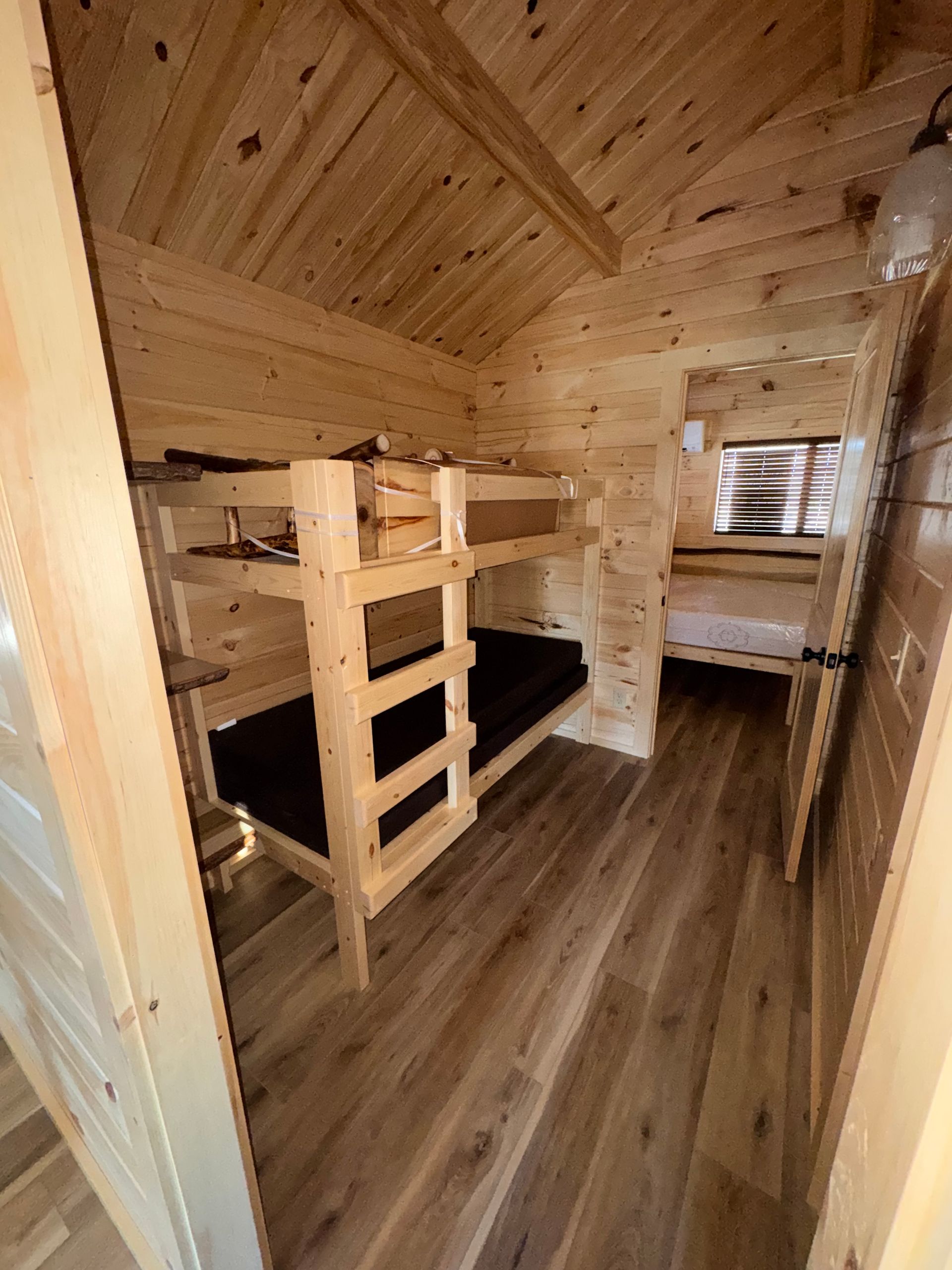 A small wooden room features a bunk bed and a doorway leading to a second bedroom with a mattress on the floor.