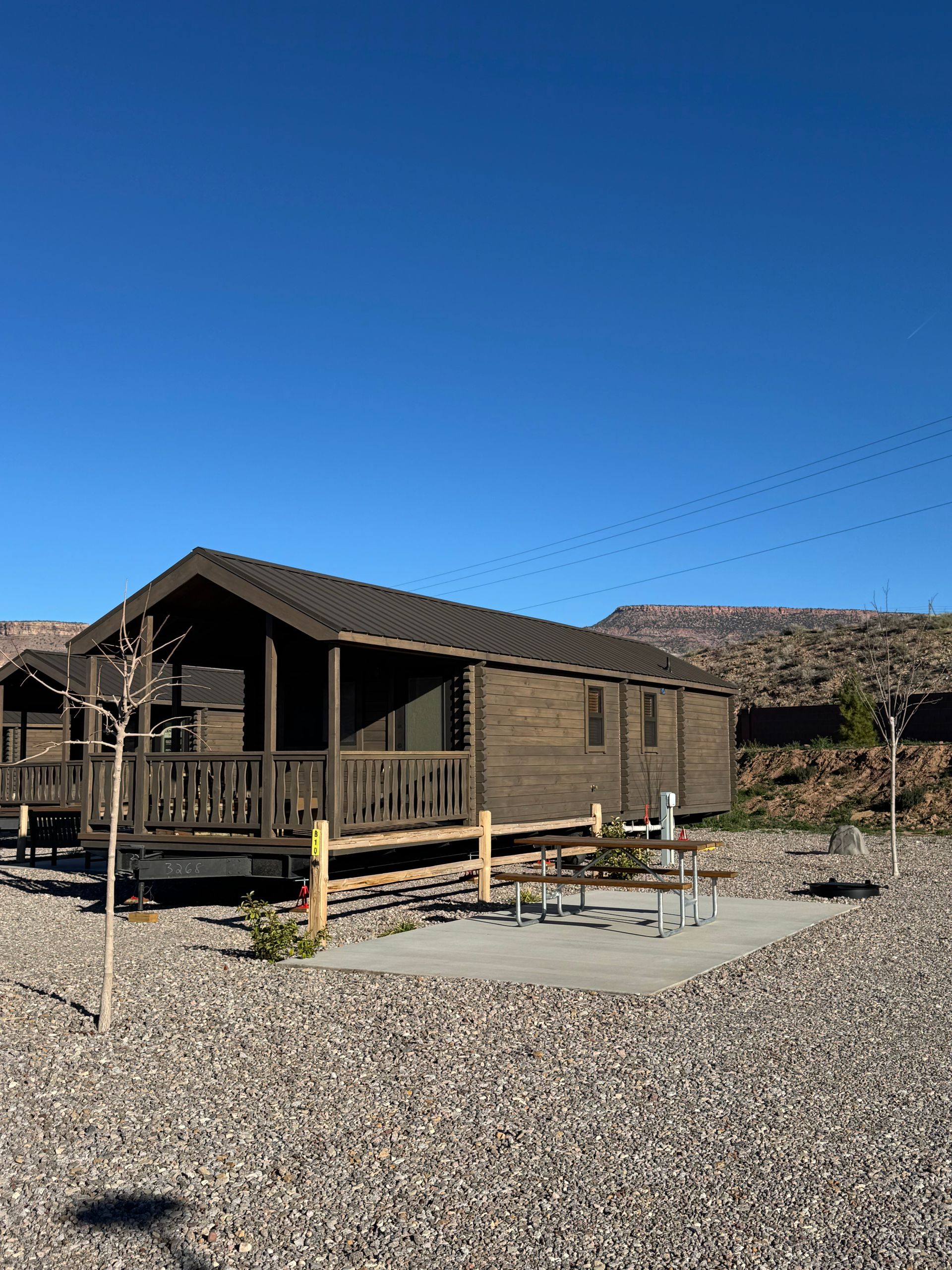 A brown wooden cabin with a porch and adjacent picnic table on a gravel lot under a clear blue sky.