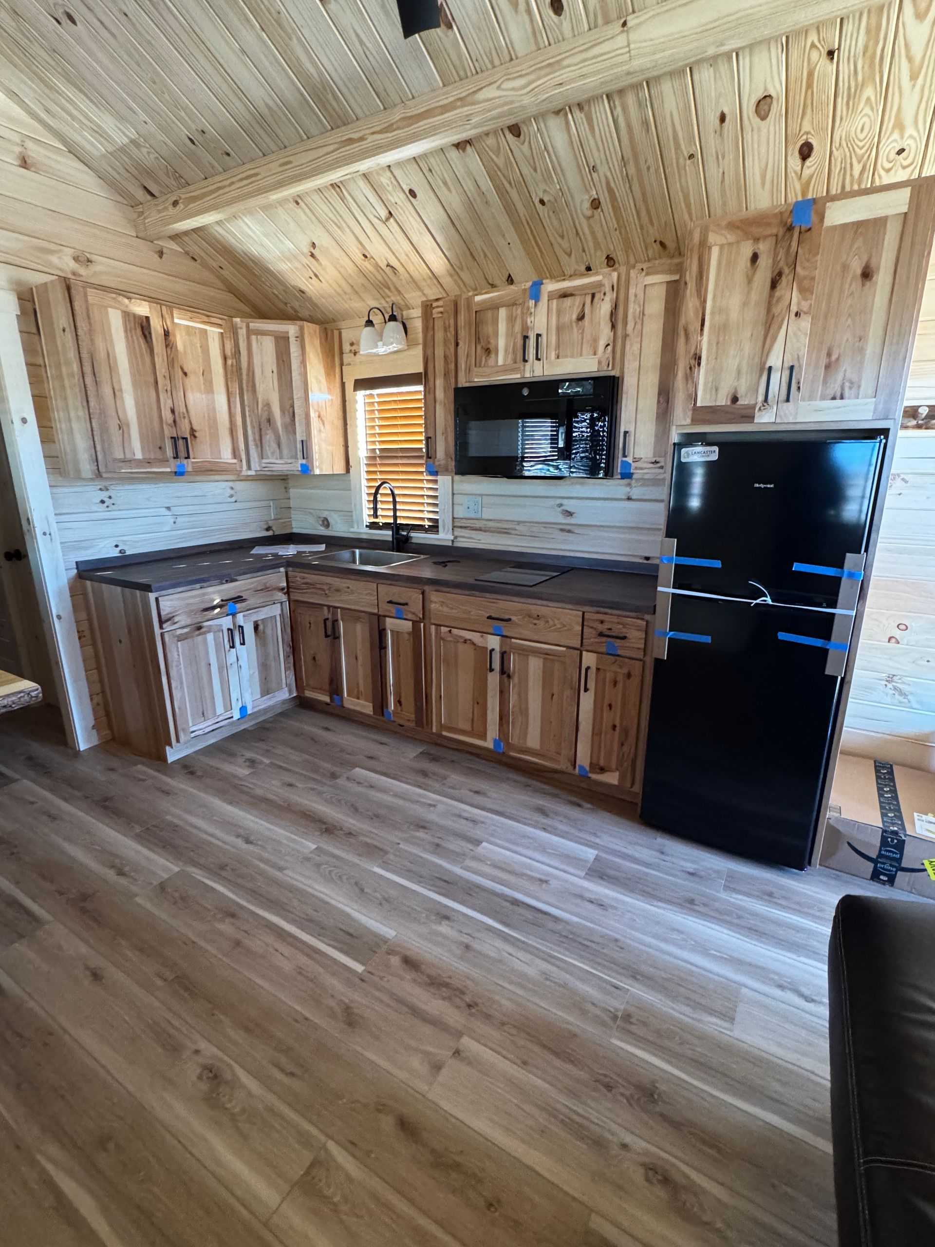 A kitchen interior with wooden cabinets, dark countertops, a black refrigerator, and wood-paneled walls and ceiling.