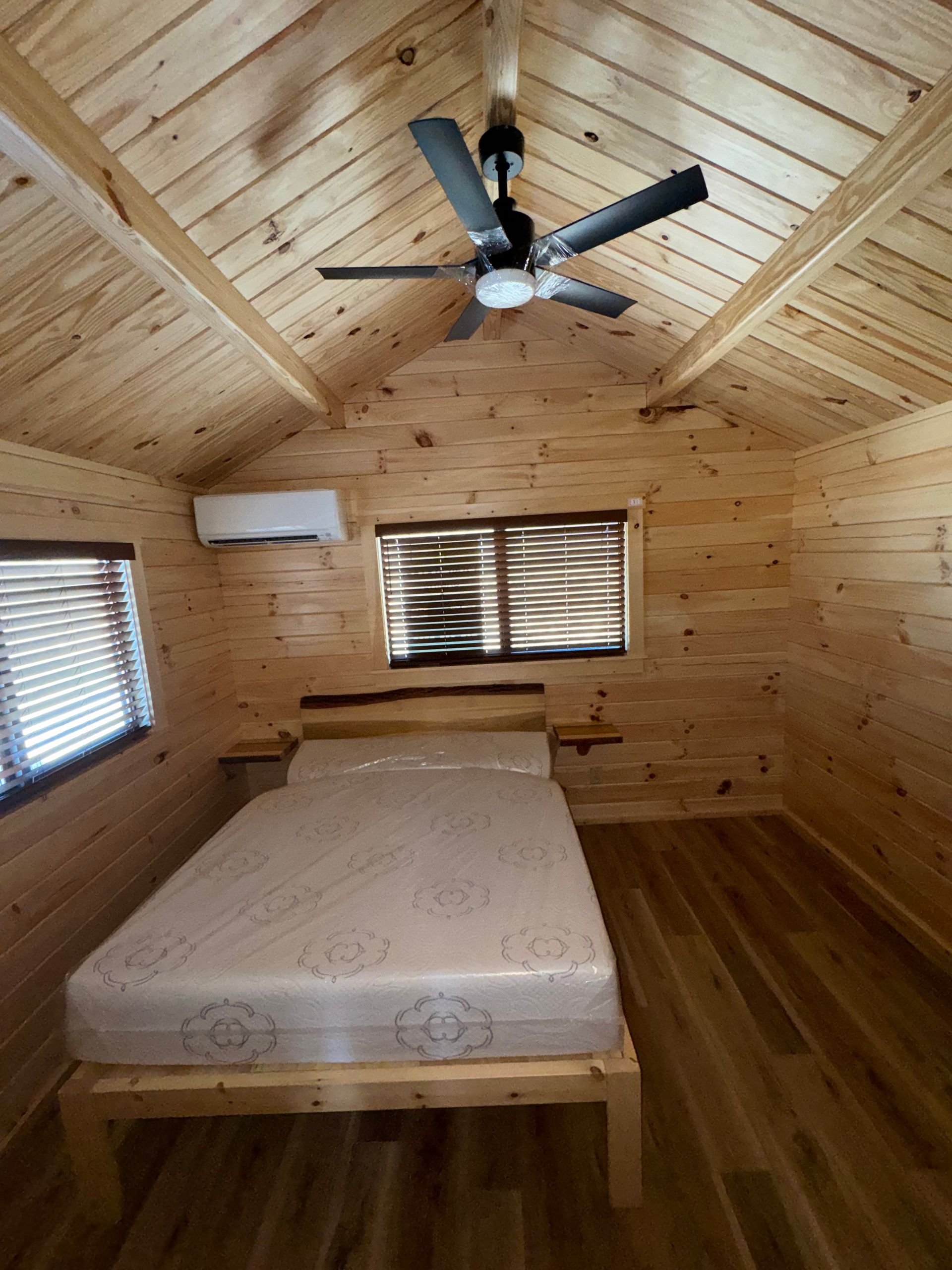 A simple cabin bedroom with wooden walls, a ceiling fan, a queen-sized mattress on a wood frame, and two windows.