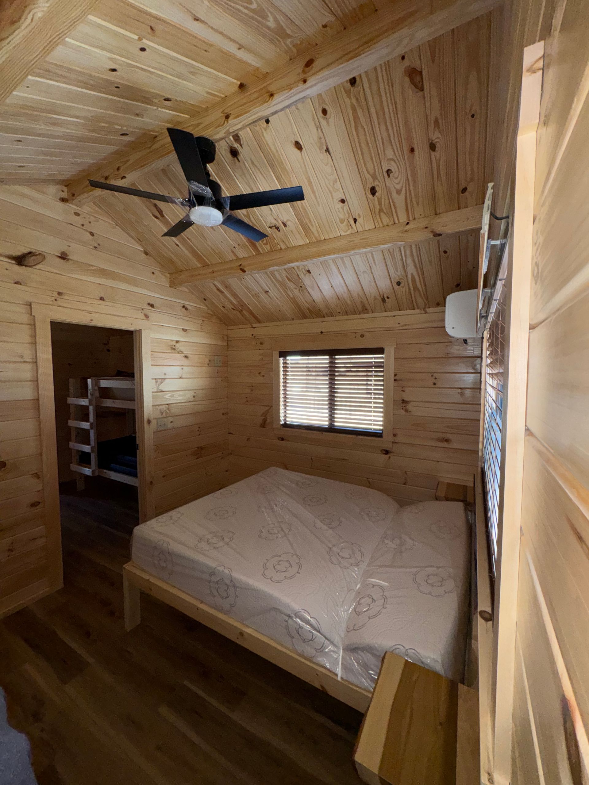 A wooden bedroom with a double bed, a ceiling fan, a window, and a glimpse into a room with bunk beds.