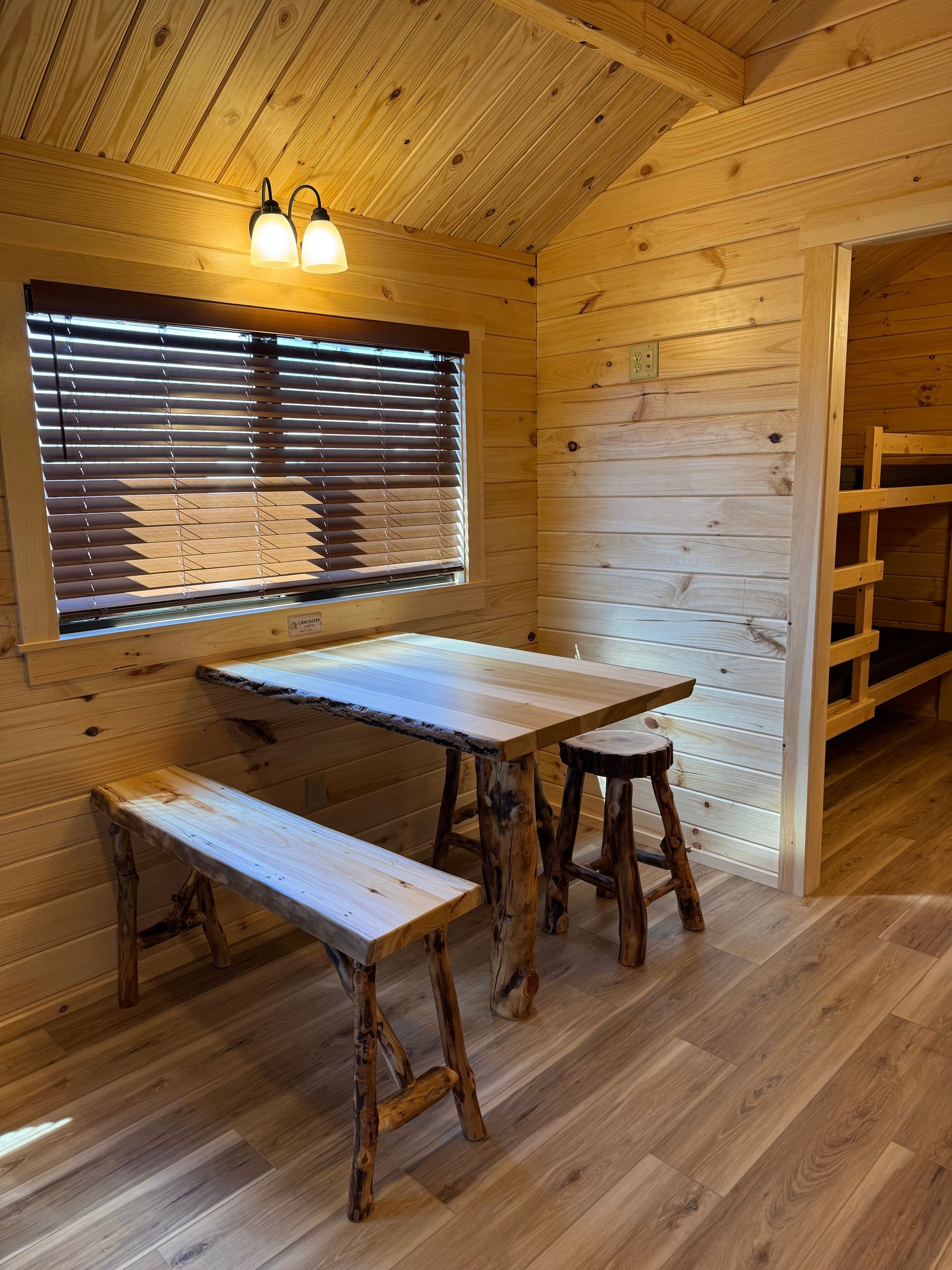 A rustic wooden cabin room featuring a wooden table with a bench and stool near a window with blinds and bunk beds nearby.