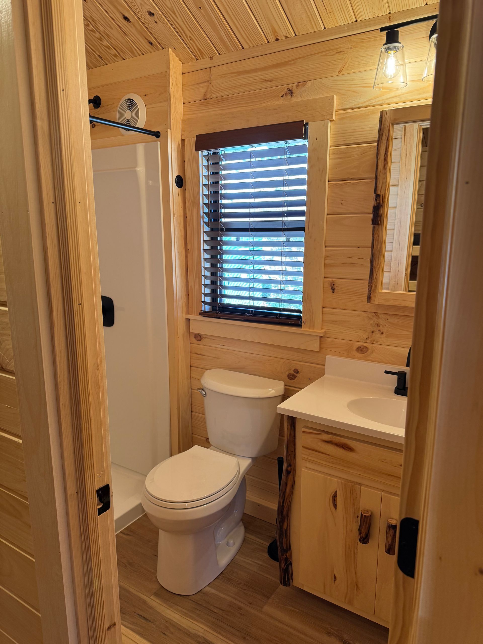 A rustic bathroom featuring light wood paneled walls, a white toilet, a wood vanity with a sink, and a walk-in shower.