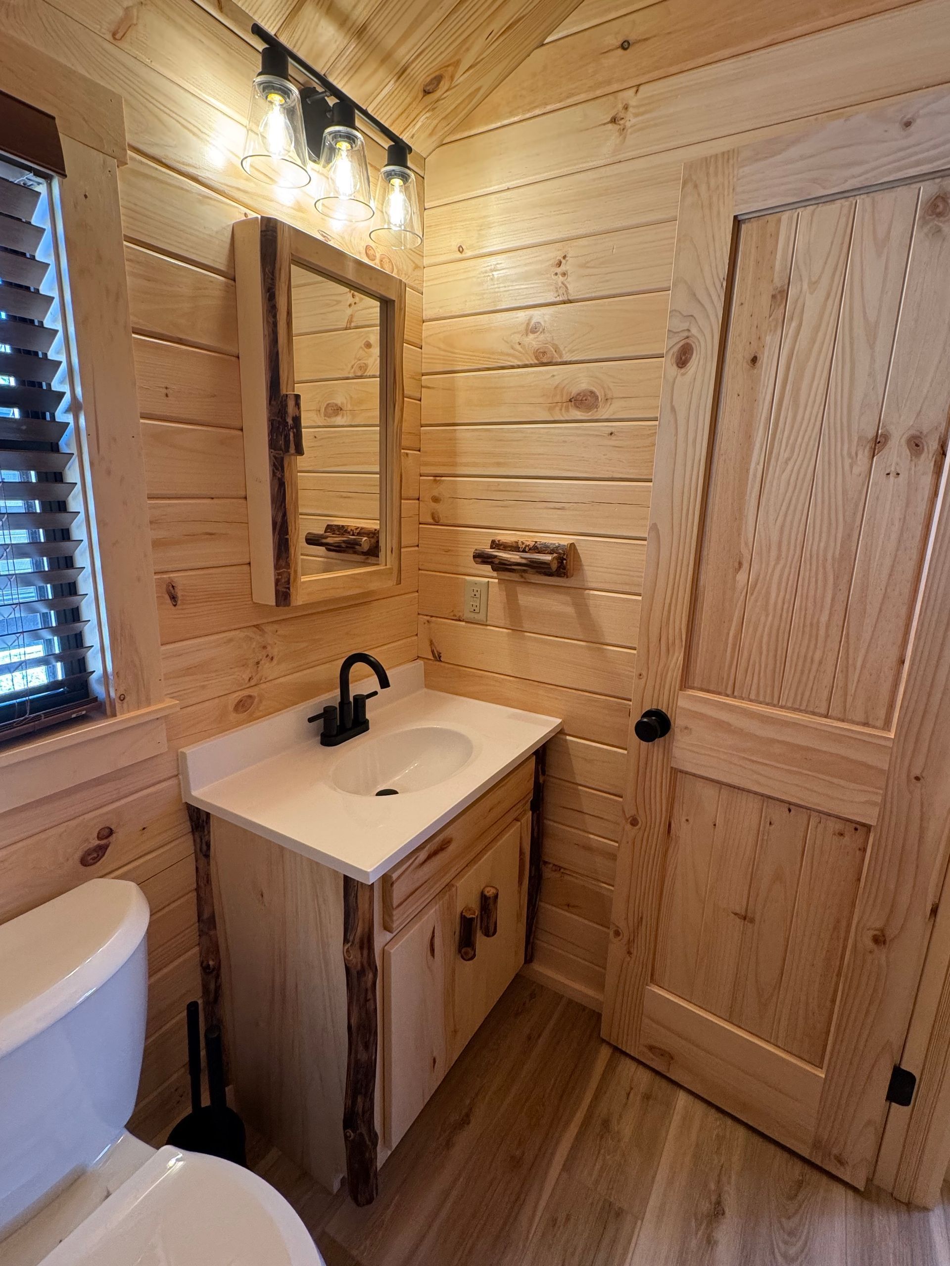 A rustic bathroom featuring light wood paneled walls, a vanity with a white countertop, a mirror, and a wood-paneled door.