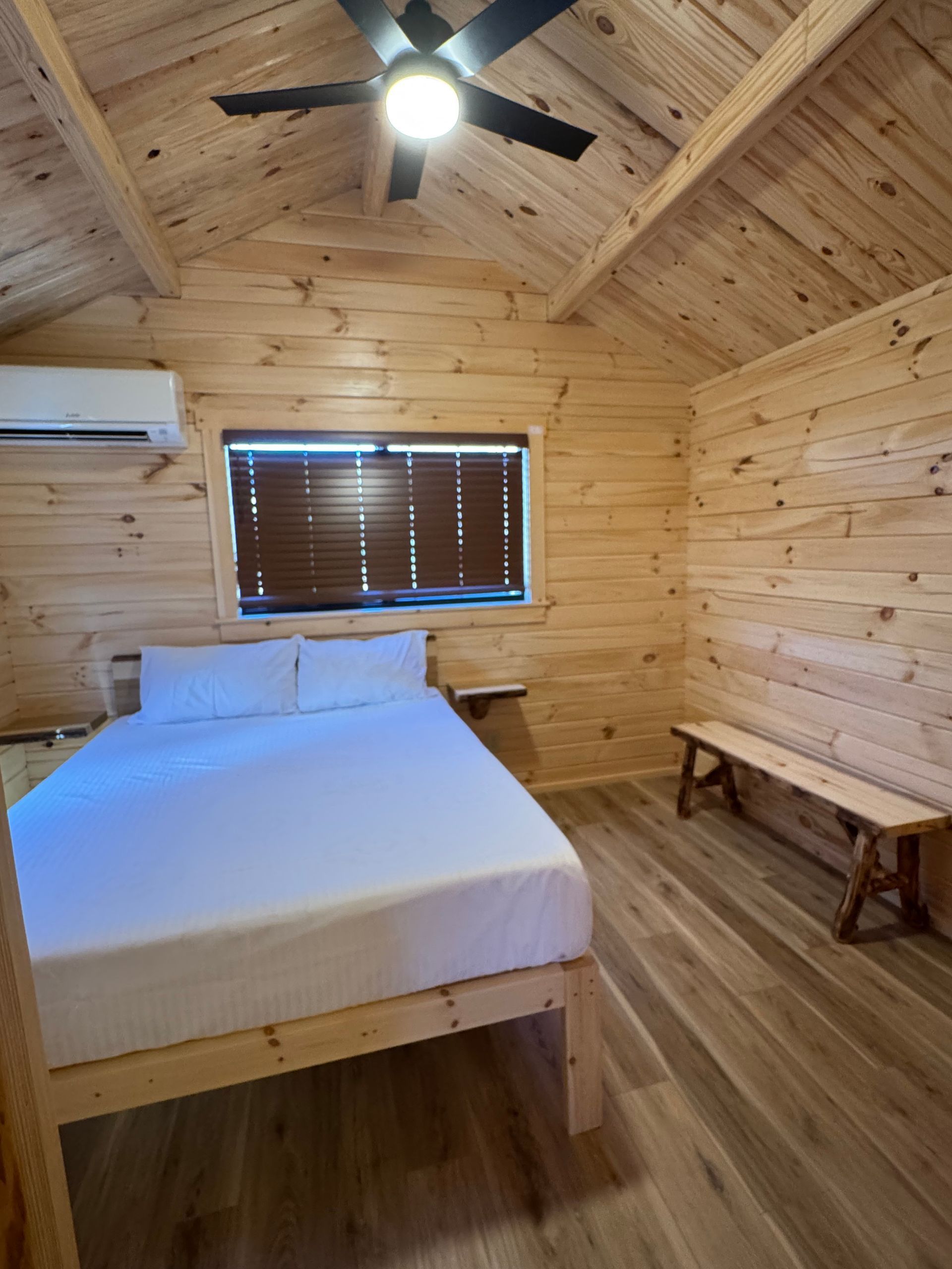 A cozy bedroom inside a wood-paneled cabin with a white bed, a wooden bench, a ceiling fan, and a window with blinds.